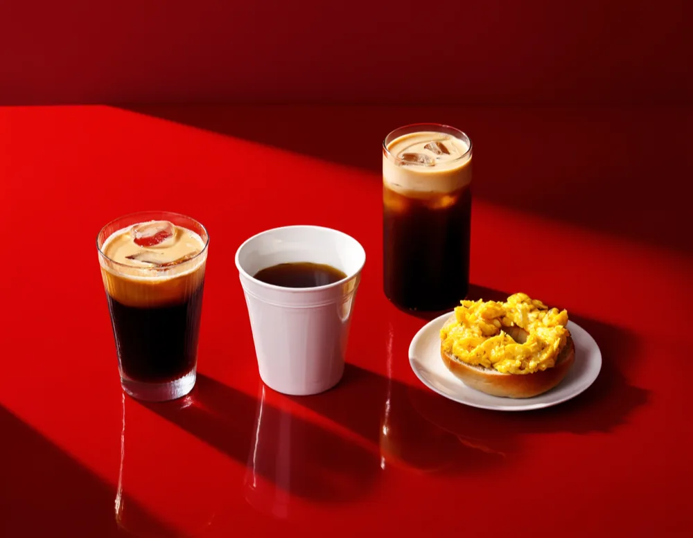 Two glasses of cold brew, a cup of filter coffee and a bagel with scrambled egg on the glossy red counter.